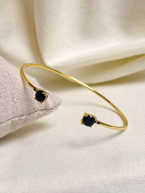 Gros plan le bracelet Nina sur un tissu blanc. Un bracelet jonc en plaqué or ouvert avec deux zirconiums noirs