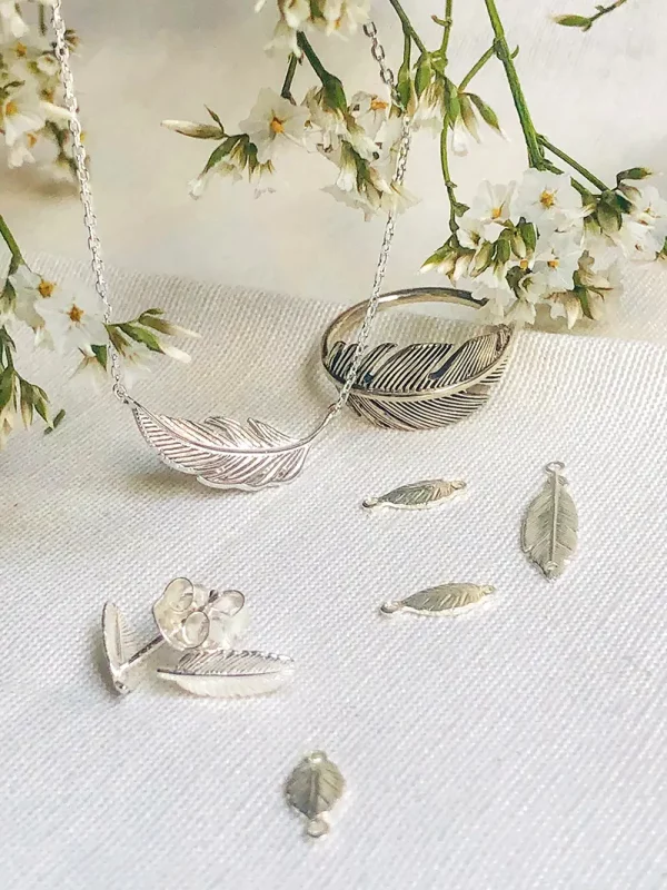 Vue de face sur un tissu avec des fleur, sont étalés des bijoux en argent avec des plumes. Le collier Plume, la bague Plume, les boucles d'oreilles Plume et le bracelet cordon Plume