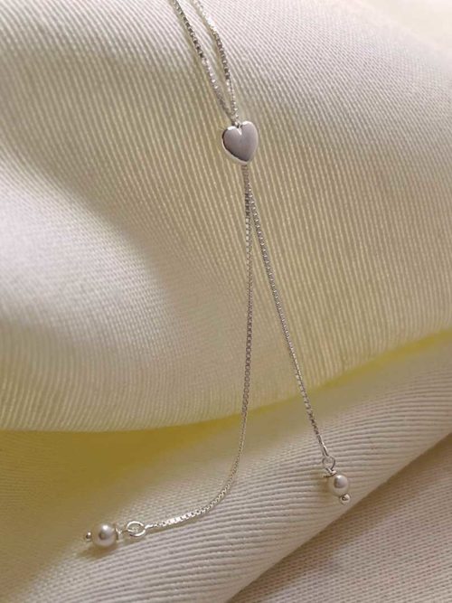 Gros plan collier posé sur un tissu blanc, un collier en argent avec un pendentif long avec deux perles tenues par un coeur