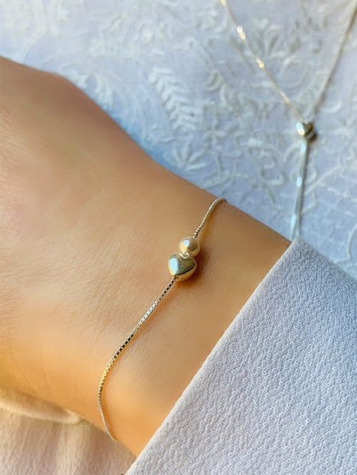 Gros plan poignet de femme avec le bracelet Eliya sur une chaîne en argent avec un coeur et une perle. Au second plan le collier Louise assorti