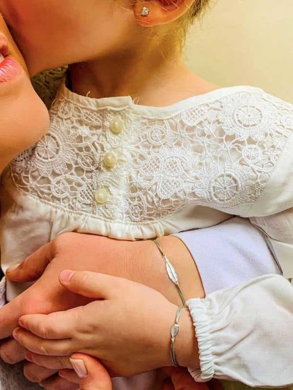 Vue de face une petite fille dans les bras de sa mère avec chacune au poignet le même bracelet. Elles portent un bracelet cordon avec une plume en argent. Bracelet cordon Plume et bracelet cordon Petite Plume