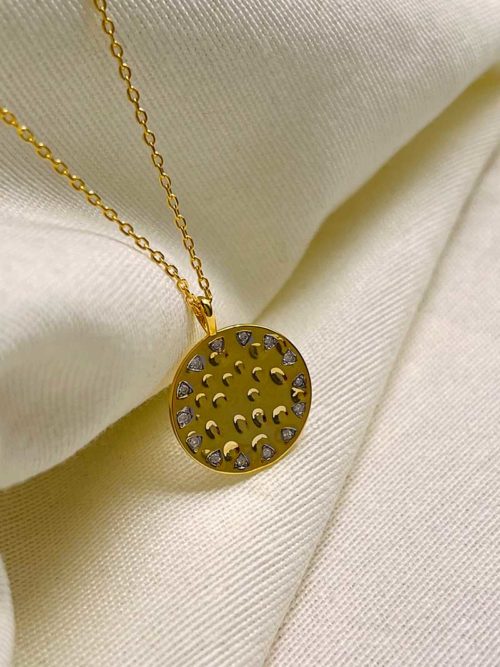 Gros plan sur un tissu blanc le collier Lya avec une chaîne et une plaque ronde en plaqué or incrustée de zircons
