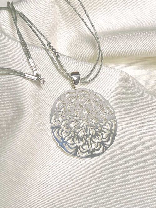 Gros plan posé sur un tissu blanc le collier cordon Mandala avec un cordon gris et un pendentif en forme de mandala en argent