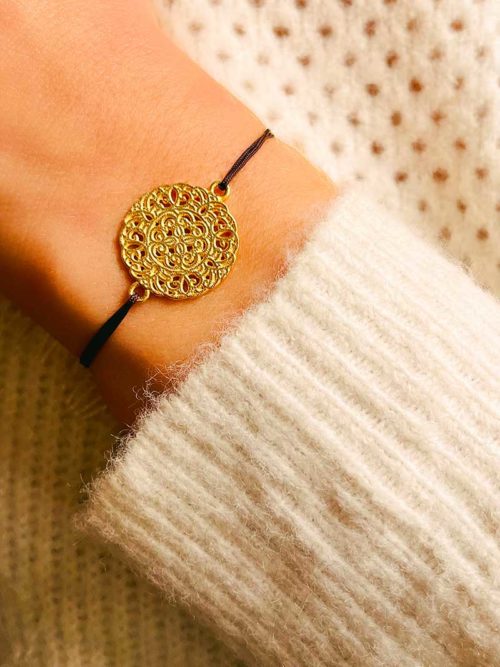 Gros plan poignet avec un bracelet cordon marron avec une plaque ronde avec des arabesque en filigrane. Bijoux en plaqué or
