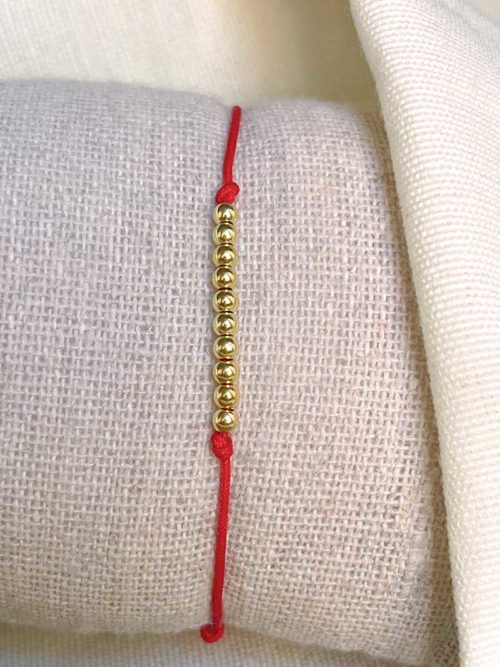 Gros plan sur un tissu blanc un bracelet cordon rouge avec une rangée de 10 perles en plaqué or