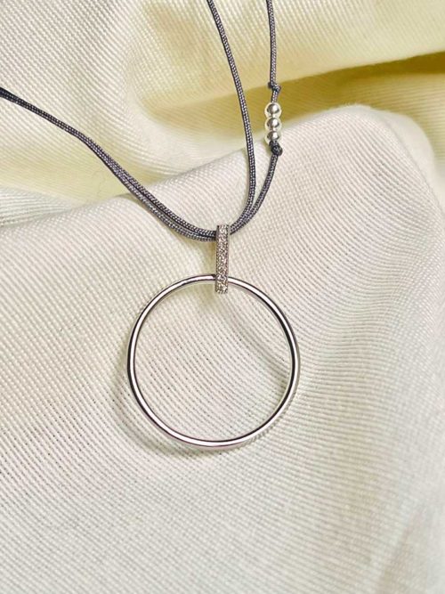 Gros plan posé sur un tissu blanc le collier cordon Manon avec un cordon gris et un pendentif en forme de cercle ajouré en argent tenu par une baguette de zircons