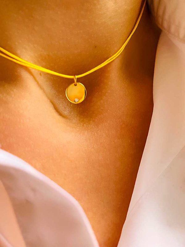 Gros plan cou avec le collier cordon Raphaëlle Doré. Il est composé d'un cordon jaune et d'une une plaque ronde en plaqué or incrusté d'un zircon
