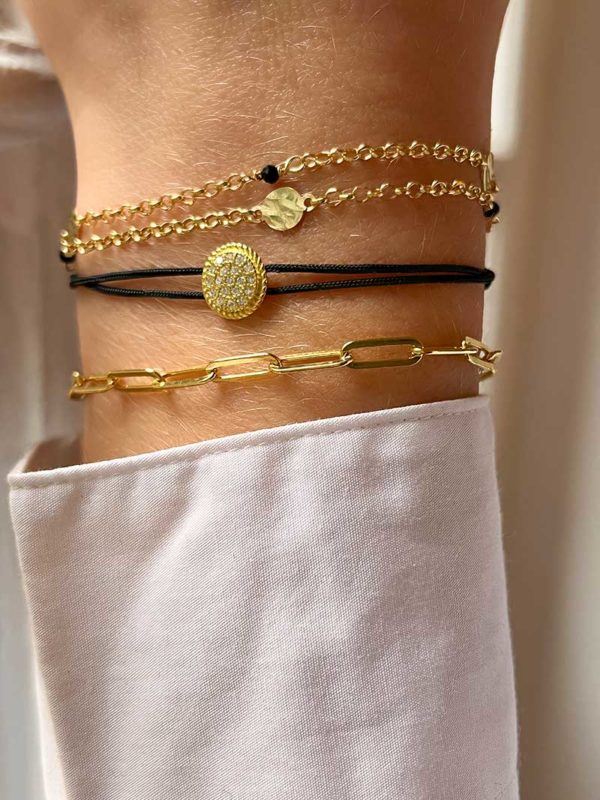 Gros plan poignet femme devant une fenêtre avec une composition de bracelets en plaqué or et cordon. Le bracelet Adèle, le bracelet cordon Alya et le bracelet Léna Doré