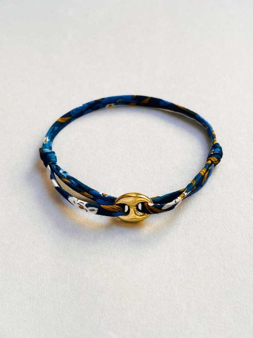 Gros plan posé devant un fond blanc le bracelet cordon Alizée doré avec une maille marine en plaqué or et un cordon imprimé liberty dans les bleus