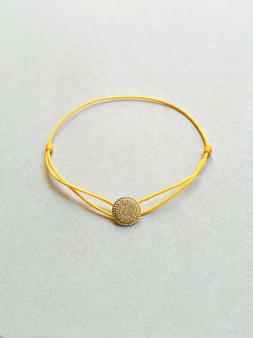 Gros plan posé devant un fond blanc le bracelet cordon Alya avec une plaque ronde en plaqué or recouverte de zircons et un cordon jaune
